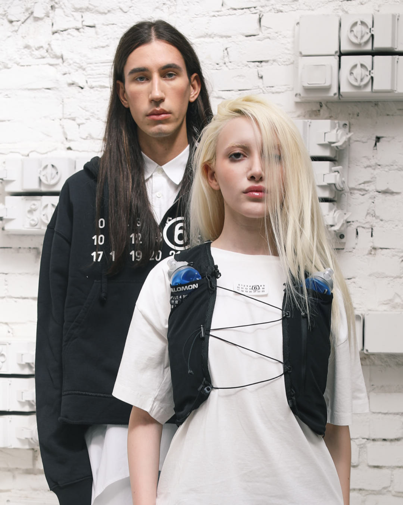 Editorial portrait for the SOCIETE ANONYME x MM6 collaboration, showing two models in layered streetwear and technical vests standing against an industrial white brick backdrop.