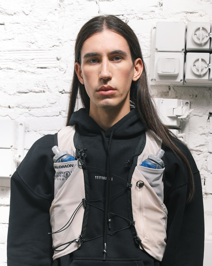 Close-up fashion image from the SOCIETE ANONYME x MM6 collaboration, featuring a model in a black hoodie and light technical vest with water bottles against a textured white brick wall.